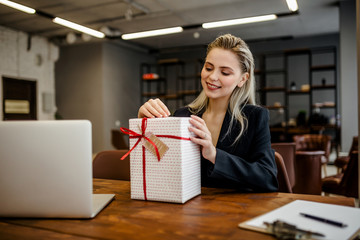 A beautiful blonde businesswoman sits at a Desk in the office and with a smile opens a gift that was given by colleagues at work