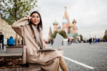 Beautiful business woman of Asian appearance in glasses working at a laptop on the background of red square in Moscow