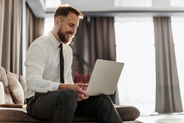 A bearded businessman in a shirt sits in the office and with a smile working on a laptop
