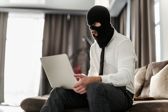 A male hacker in a mask and shirt is working on a laptop, trying to hack the company's management system