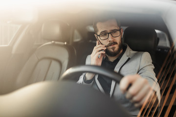 A bearded businessman with glasses is sitting behind the wheel of an Executive car and talking on the phone. Photo through the windshield