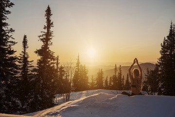 Young female wearing long hair dreads, seating in yoga lotus pose and meditating on sunrise in snow forest on mountain top. Cold frozen winter morning