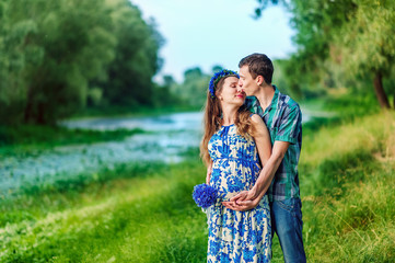 Happy and young pregnant couple hugging and kissing in nature with bouquet of knapweeds in hands. New family expecting baby in scenic place near river in summertime. Happiness to be parents.