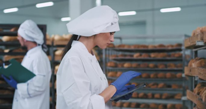 Good looking lady with a new big tablet take some photos of a fresh baked bread from the shelves in a big bakery factory in the background two other bakers chatting. 4k