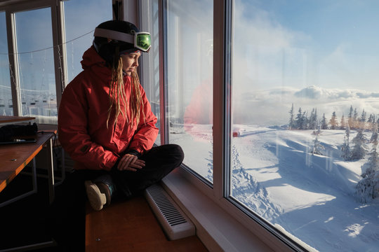 Woman snowboarder sitting in cafe enjoying and relaxing. Window with panoramic view to winter snowy mountain slop in ski resort