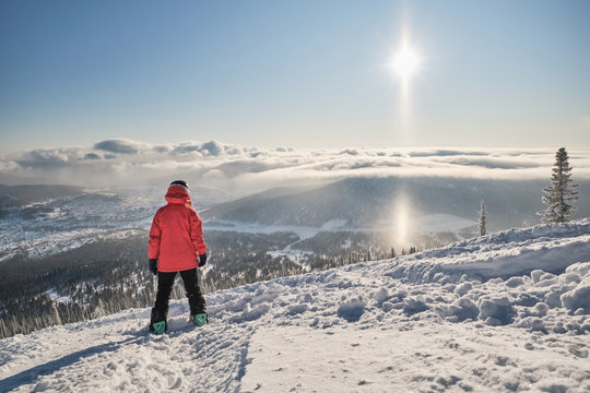 Female snowboarder standing on mountain slop above clouds and beautiful mountains landscape, preparing to snowboarding. Sunny winter day in ski resort