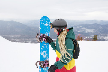 Woman snowboarder wearing dreadlocks and rasta colours hoody standing on mountain top, preparing to...