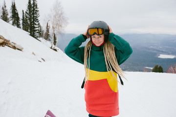 Woman snowboarder wearing dreadlocks and rasta colours hoody standing on mountain top, preparing to...