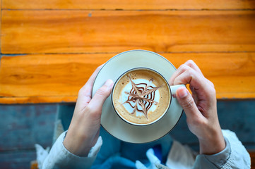 Woman's hands in sweater hold a cup of strong coffee on wooden table. Coffee fan top view...