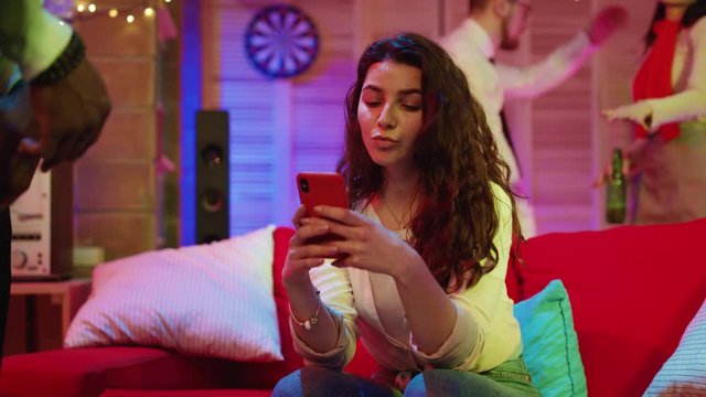 Beautiful woman with curly black hair sitting on sofa browsing smartphone device chilling out at best friends night party.