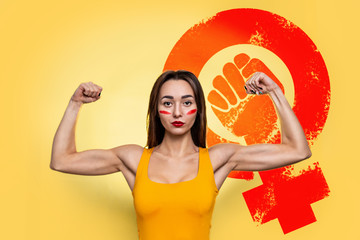 Feminism and the concept of equal rights and independence for women. A Caucasian young woman in an orange t-shirt shows off her pumped-up biceps. Yellow background. Copy space. Sign of girlpower