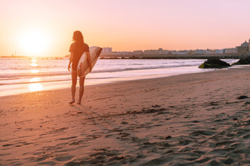 Fototapeta na wymiar against the background of the sun setting over the horizon, a girl with a surfboard goes to the ocean water