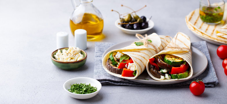 Wrap sandwich with grilled vegetables and feta cheese on a plate. Grey background.