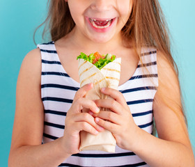 Happy beautiful child girl eating wrap sandwich. Blue background. Close up.