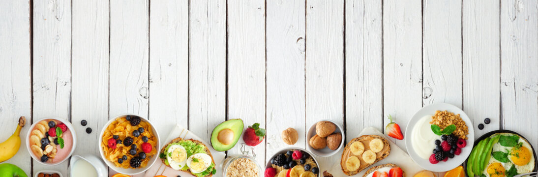 Healthy breakfast food banner with bottom border. Table scene with fruit, yogurt, smoothie bowl, nutritious toasts, cereal and egg skillet. Top view over a white wood background. Copy space.