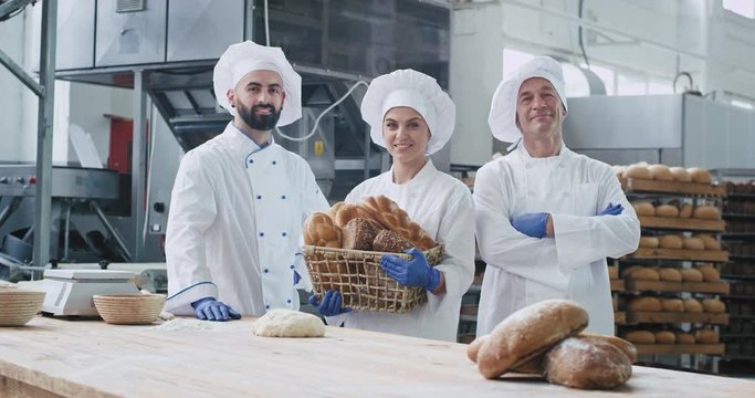Two generations of bakers two men and woman in a big bakery industry looking straight to the camera and smiling beside their table work while holding a basket of organic bread