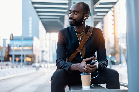Young businessman with coffee to go listening music with earphones and smartphone relaxing at tram stop