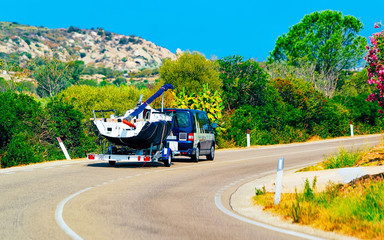 Car trailer with yacht or motorboat on road at in Sardinia Island, Italy summer. Minivan with motor...