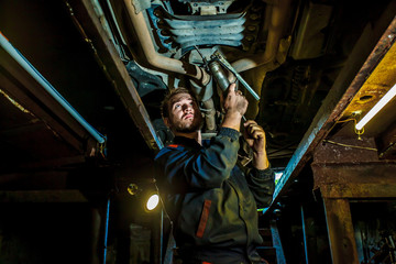 Fototapeta na wymiar Handsome mechanic in uniform are working in auto service with lifted vehicle. Car repair and maintenance. Mechanic examining under the car at the repair garage. 