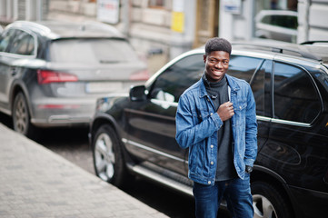African man wear on jeans jacket posed outdoor, standing against black car.