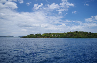 A small forested island in the Philippines archipelago. Isle is surrounded by the blue, calm waters of the ocean and azure, clear sky. There are few clouds, day is very bright and sunny. 