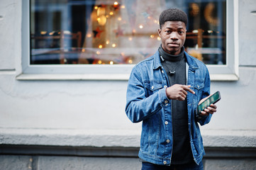 African man wear on jeans jacket posed outdoor with cell phone at hand.