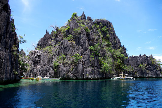 Big rocky island in the Philippines archipelago. Two paraw boats (double outrigger sail ships) are sailing near. The water of the ocean is calm and rippling slightly, it's a clear summer day.