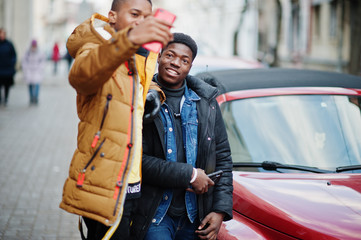 Two african male friends standing near red american convertible car and making selfie on phone.