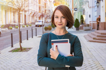 A young student with textbooks in her hands, looking at the camera on a city street on a sunny day....