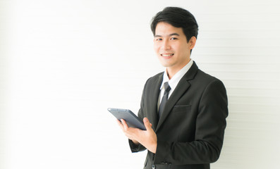 Young Asian handsome businessman wearing black suit smiling and holding tablet looking camera standing on white background with copy space