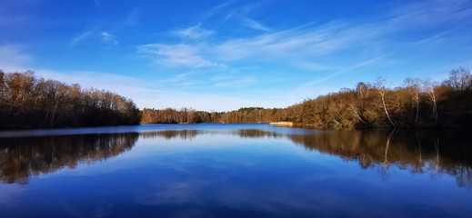 Naklejka na ściany i meble A quiet calm lake in January with different dark and light blue tones sky and trees that are reflected on the shore in the water and magnificent panorama with far-sightedness