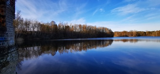 A quiet calm lake in January with different dark and light blue tones sky and trees that are reflected on the shore in the water and magnificent panorama with far-sightedness