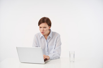 Shot of confused young short haired brunette lady with casual hairstyle looking at screen of her laptop with displeased face, reading unpleasant news, isolated over white background