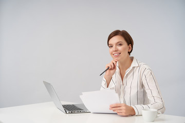 Cheerful young attractive brown-eyed short haired woman smiling pleasantly while looking at camera and leaning chin on raised hand, posing over white background with laptop and documents