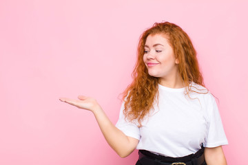 young red head woman feeling happy and smiling casually, looking to an object or concept held on...