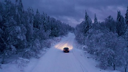 Aerial: flying above car driving alone through a snowy frozen forest at cloudy dark evening. Empty rural road surrounded by snow covered trees. - Powered by Adobe