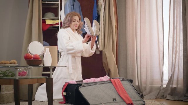 Young female Caucasian tourist putting stuff into suitcase and closing bag. Tired cheerful woman packing for a trip at home. Lifestyle, leisure, tourism.