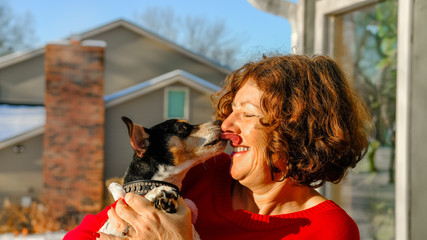 Smiling attractive mature woman in red sweater holding small dog; dog licking woman's face
