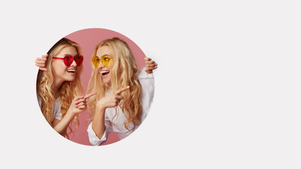 Portrait of two young happy, shocked women in the heart shape sunglasses looking throth the white hole in the wall. Big sale. Funny faces. Empty space for text