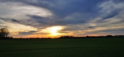 A fantastic sunset with red yellow orange and purple tones with dramatic cloud formation over green meadows with bare trees in January on the island of R&uuml;gen