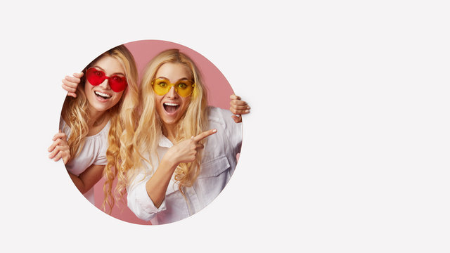 Portrait of two young happy, shocked women in the heart shape sunglasses looking throth the white hole in the wall. Big sale. Funny faces. Empty space for text