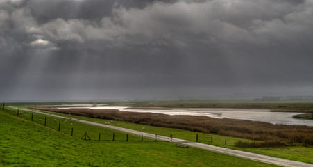 Sunrays in a dark sky above the coastal landscape in the northern Netherlands; a dike and wetlands