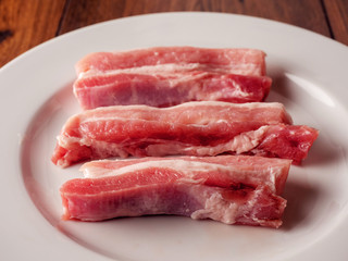 Uncooked raw pork belly slices on a white plate and wooden table.