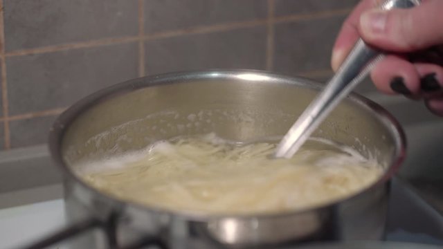 cook hand stirs spaghetti in silver pot with boiling water using spoon on white stove in kitchen close view
