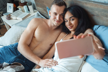 Mixed Race Couple. Young man and woman at home lying on bed taking happy selfie on smartphone