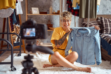 Fashion Blogger. Young woman influencer in glasses sitting on carpet at stylish apartment recording vlog on camera with denim shirt laughing cheerful