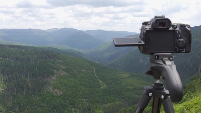A camera on a tripod in nature overlooks view of valley, nature and mountains on a cloudy day.