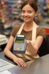 Daily Shopping. Young cashier woman at the supermarket offering credit card reader close-up blurred background smiling joyful