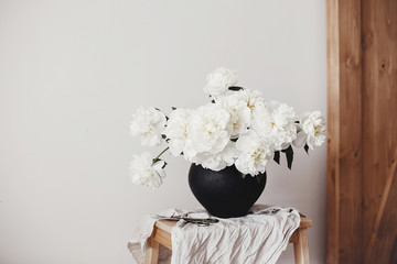  White peonies rural still life. Stylish peony bouquet in black clay vase on linen fabric with...