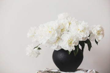 Stylish peony bouquet in black clay vase on linen fabric with scissors on rustic background. White peonies rural still life. Hello spring wallpaper. Happy Mothers day. Space text